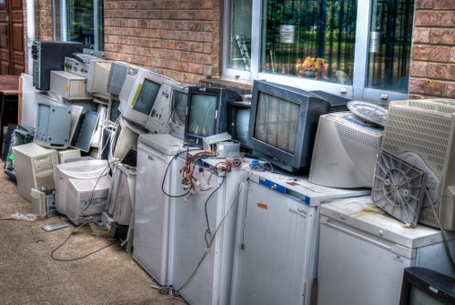 A row of discarded electronic waste, including various outdated computer monitors, CRT screens, and tower units, arranged on a paved outdoor surface next to a brick building with large windows. The monitors display different shades of grey and black screens, with some showing visible cables and connectors hanging loosely. The electronic appliances appear aged and decommissioned, with some units showing signs of wear such as faded surfaces or missing parts. Behind the appliances, the brick wall of the building and a window with reflections of greenery can be seen, indicating a suburban or urban street setting. The image represents a collection of obsolete electrical equipment waiting for proper rubbish removal and recycling, illustrating the importance of responsible waste disposal services like those offered by Mayfair House Clearance in the London area, which includes handling electronic waste responsibly. The overall scene has neutral natural lighting, emphasizing the array of different sized and shaped electronic items, contributing to an accurate visual depiction suitable for a rubbish removal service website.