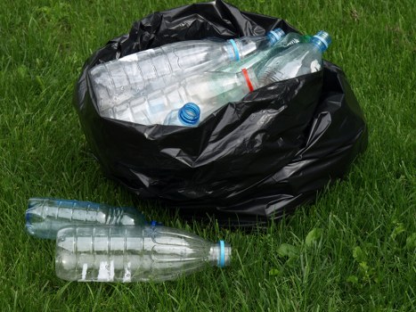 The image shows a large black garbage bag placed on a grassy area, filled with several clear plastic water bottles with blue caps and labels, some of which are partially protruding from the top of the bag. Two additional empty plastic bottles with blue caps are lying on the grass in front of the bag. The bottles vary in size but generally appear to be standard single-use beverage containers, with some showing slight crinkling or compression. The environment appears to be an outdoor setting with fresh green grass and natural daylight, suggesting a typical scene where rubbish might be gathered for collection or disposal. This visual aligns with rubbish removal services, such as those offered by Mayfair House Clearance, particularly for waste like plastic bottles and general outdoor rubbish, with the setting hinting at a residential or localized area within London, possibly in Westminster or nearby areas. The scene underscores the need for proper waste collection and disposal services, supporting local rubbish clearance operations.