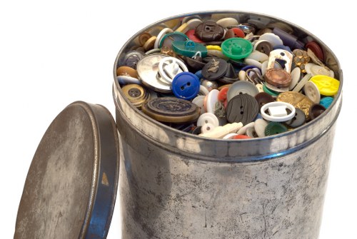 A large cylindrical metal bin filled with a variety of loose plastic and metal clothing buttons, including different sizes, shapes, and colours such as black, white, blue, green, and brown. The surface of the bin shows signs of wear and oxidation, with some areas appearing rusty and weathered. The lid, made of similar metal, rests partially open and leaning against the bin's side, revealing the densely packed assortment inside. The background is plain and neutral, highlighting the focus on the bin and its contents, which are associated with textile or clothing waste recycling or disposal services in the UK, possibly around Mayfair or central London as inferred from the webpage context of Mayfair House Clearance Services. This detailed visual description supports the context of rubbish removal and waste management services offered by Mayfair House Clearance, including handling of small waste items such as clothing accessories like buttons.